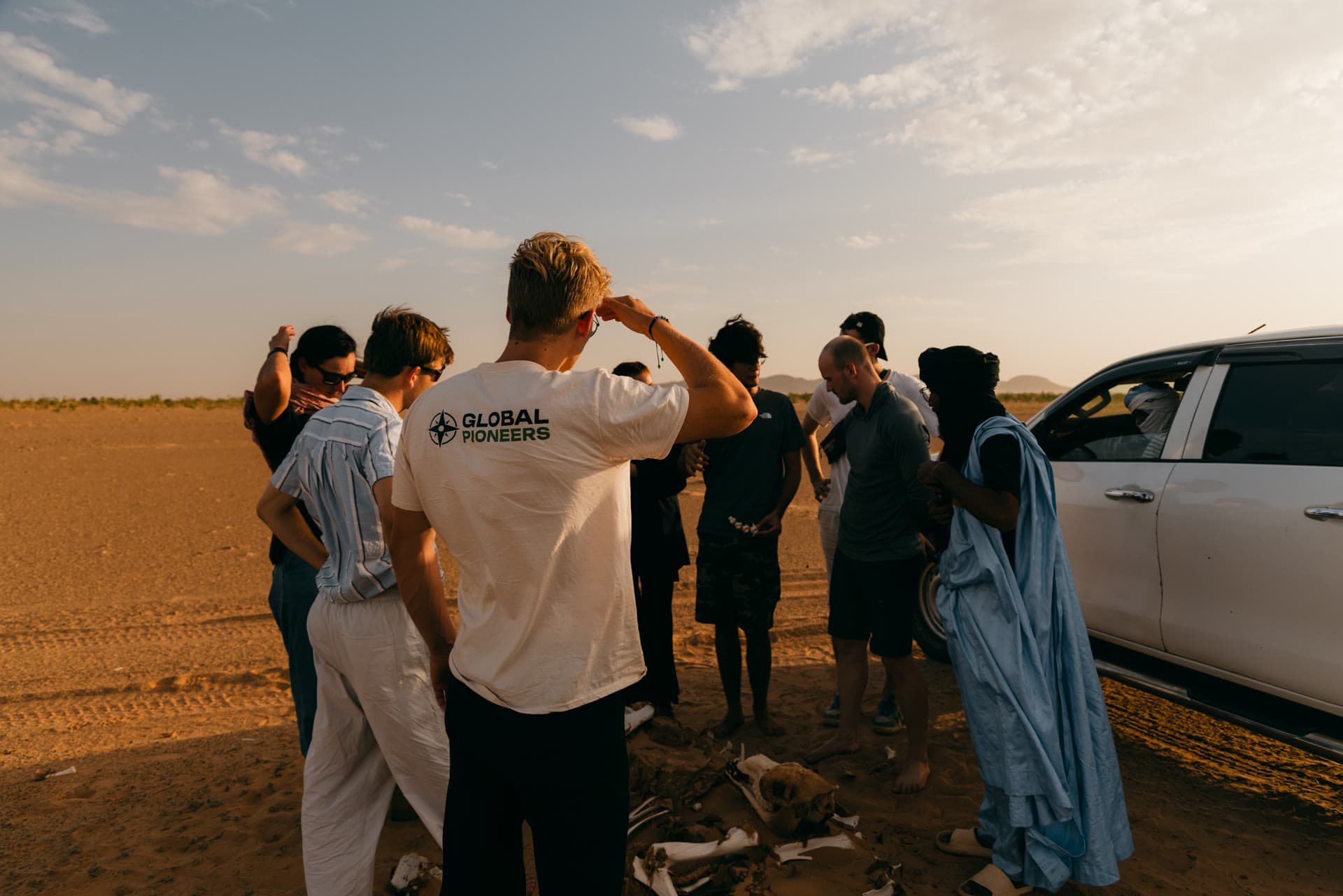 Reisegruppe und lokaler Guide stehen bei Sonnenuntergang in der Wüste neben einem Geländewagen