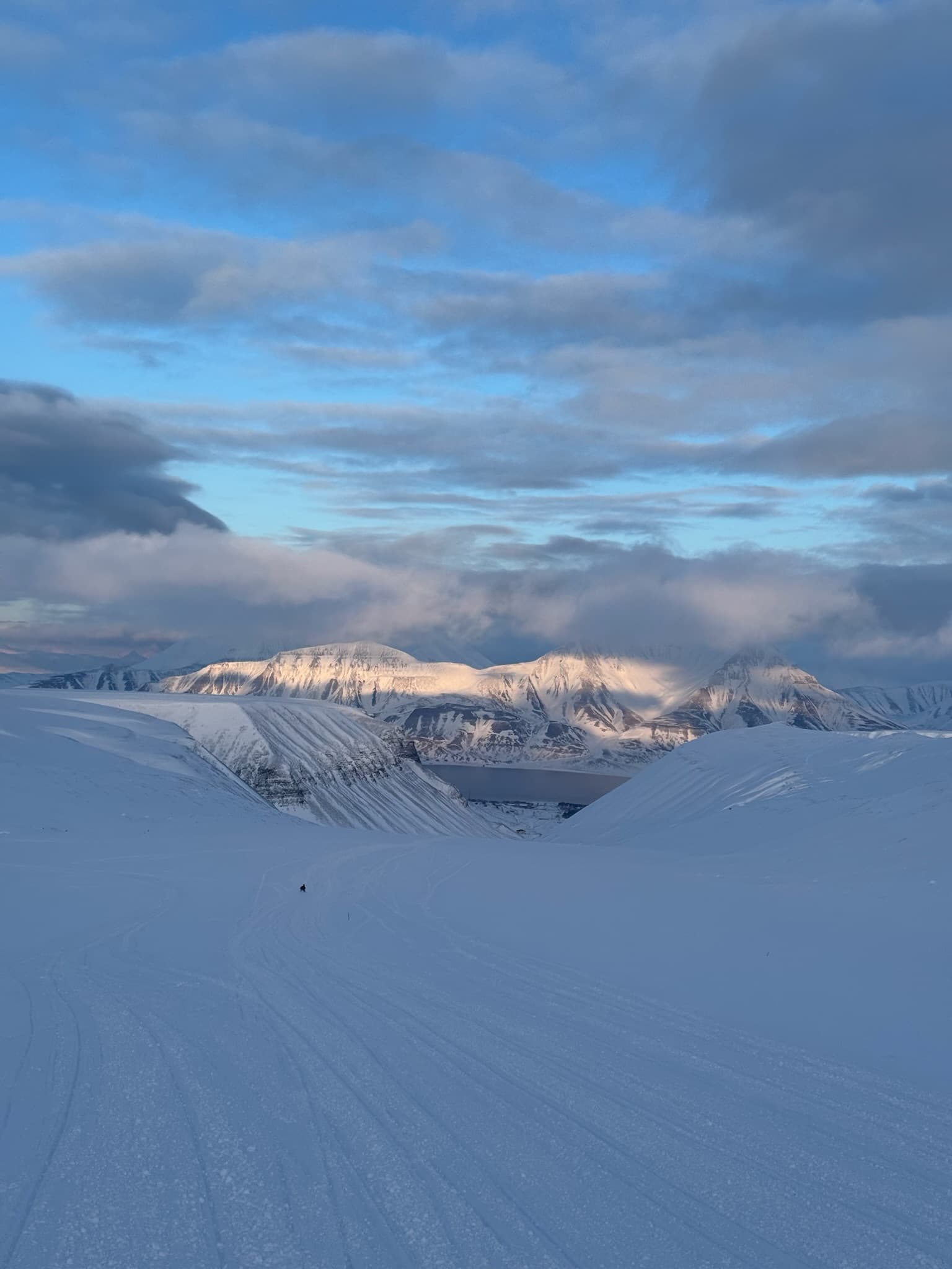 Schneemobilfahrer als winziger Punkt vor golden beleuchteten Bergketten auf Spitzbergen in der Abenddämmerung
